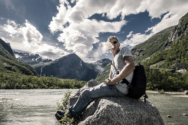 Man blowing smoke out of his mouth and sitting on a rock in front of a lake and mountains